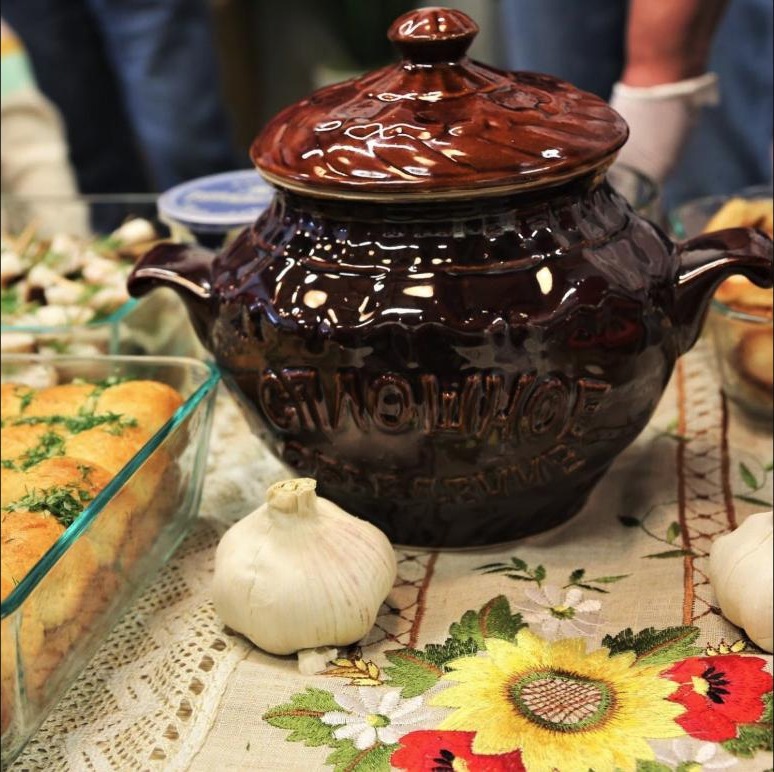 Dark ceramic borsch pot on a patterned tablecloth, surrounded by garlic and fresh bread — from a past Borsch Competition.